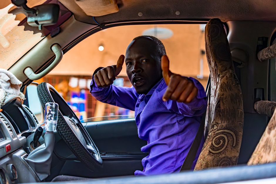 A cheerful taxi driver wearing a purple shirt gives a thumbs up inside his car, creating a warm and welcoming vibe.